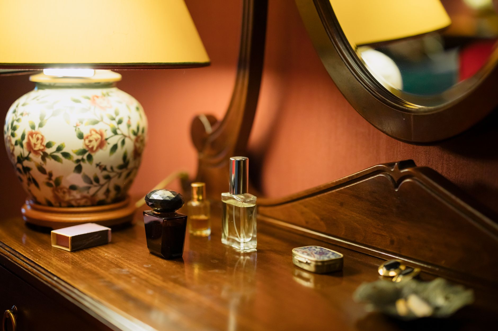 Flacons de parfum, petite boîte décorée et boîte d’allumettes sur une coiffeuse en bois, avec une lampe fleurie et un miroir ovale.