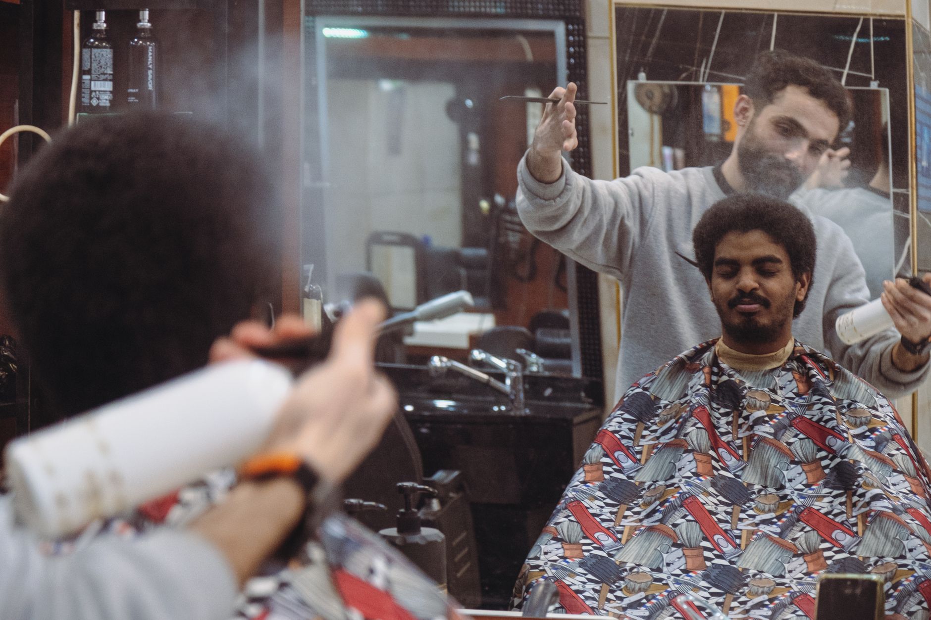 Un coiffeur vaporise un spray sur la chevelure d’un client assis sous une cape dans un salon de coiffure.