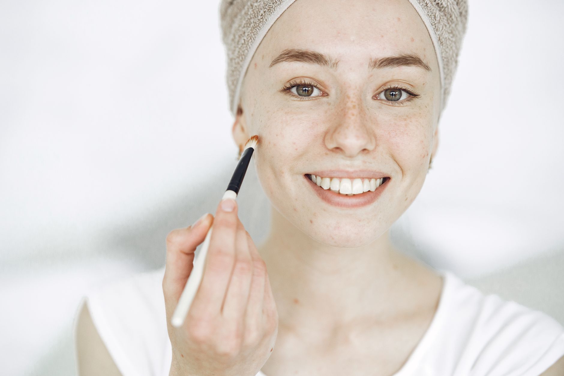 Une jeune femme souriante appliquant un produit sur sa joue avec un pinceau fin.